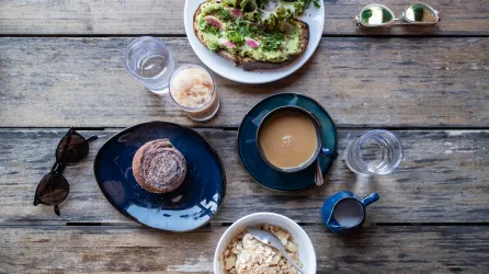 A breakfast is spread across a table with a coffee, bowl of oats, cinnamon roll, water, and avocado toast. There are other props on the table including sunglasses and water cups.