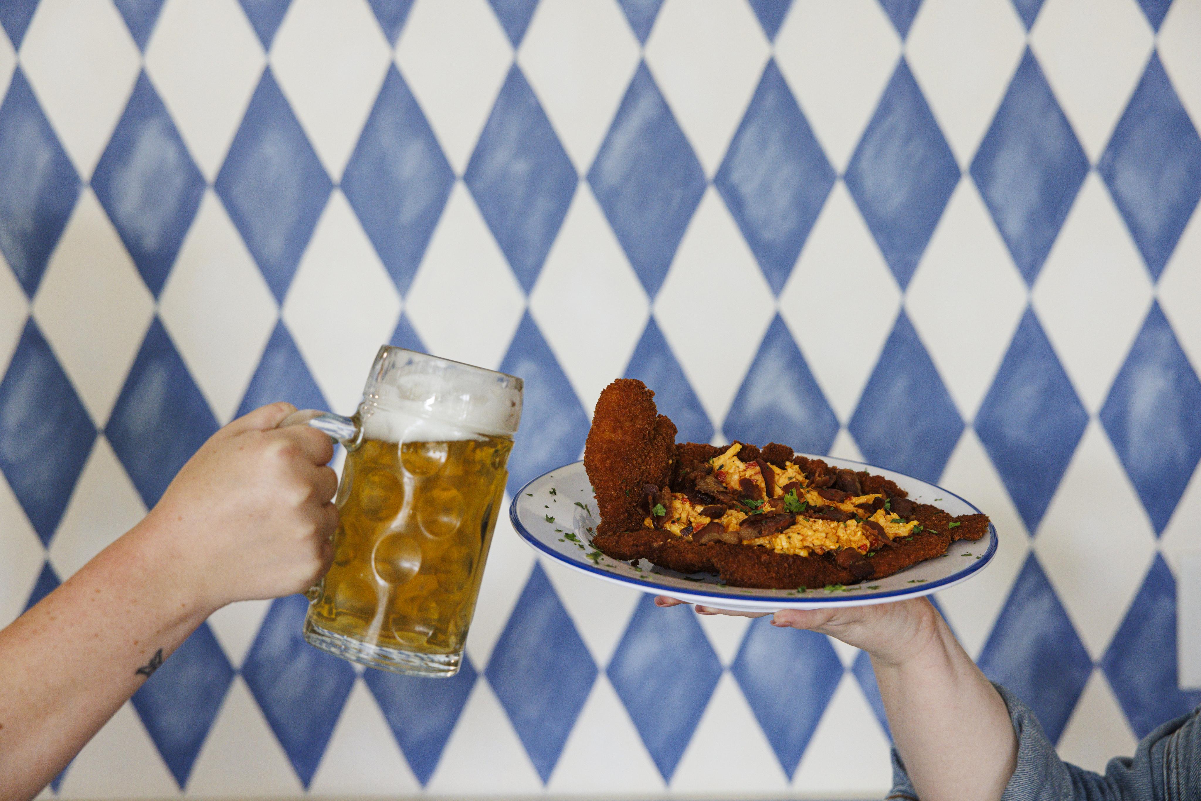 Someone is holding a stein filled with beer on the left while someone else holds up a dish that is fried and has melted pimento cheese on top. The background is a repetitive pattern of blue and white diamonds.